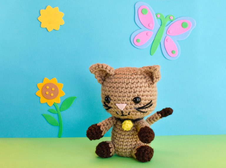 a crochet kitten doll against a blue and green backdrop with a cartoon sunflower, sun, and butterfly