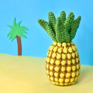 crochet pineapple against a yellow and blue fake beach backdrop with a cartoon palm tree