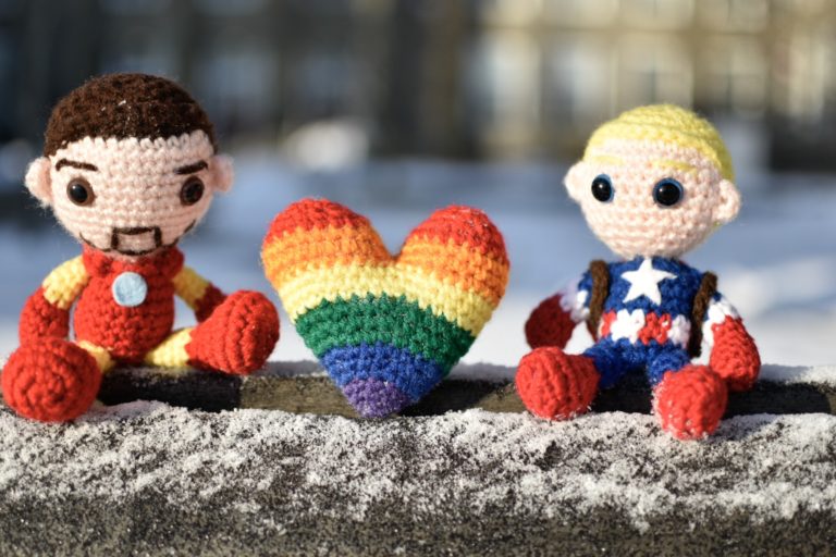 picture of crochet iron man and captain america dolls with a crochet pride flag heart between them