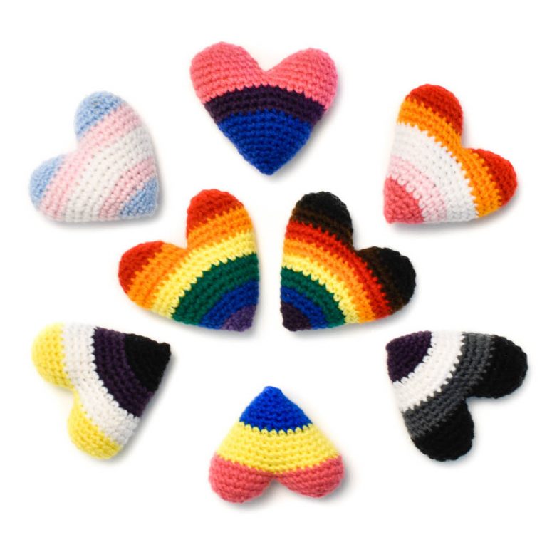 8 crochet hearts with pride colours. In the middle are two hearts in the rainbow Pride & Inclusive Pride flag colours, and around them are hearts depicting the Trans, Bisexual, Lesbian, Asexual, Pansexual, and Non-Binary flag colours.
