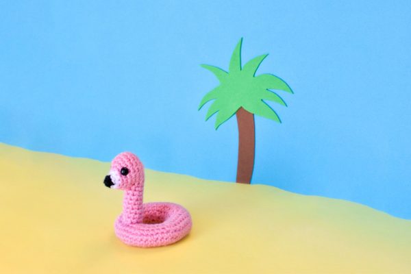 a small crochet flamingo floatie toy against a fake tropical beach backdrop made out of construction paper