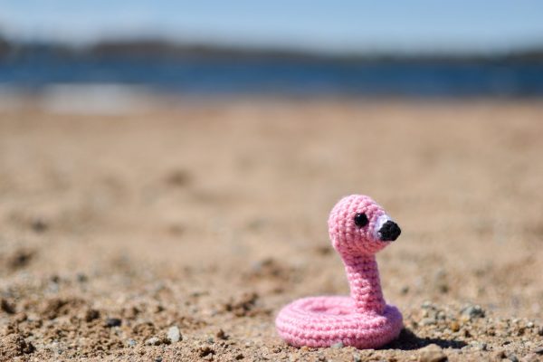 a small crochet flamingo floatie toy sitting on a beach in front of a lake