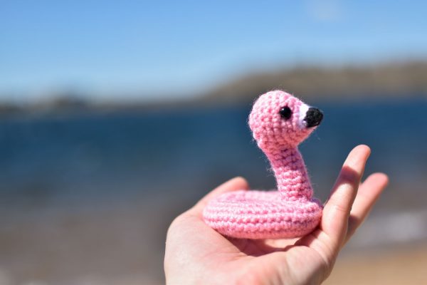 a small crochet flamingo floatie toy held up in front of a lake beach