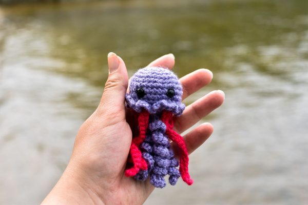 a small crochet purple and pink jellyfish toy held up above ocean water