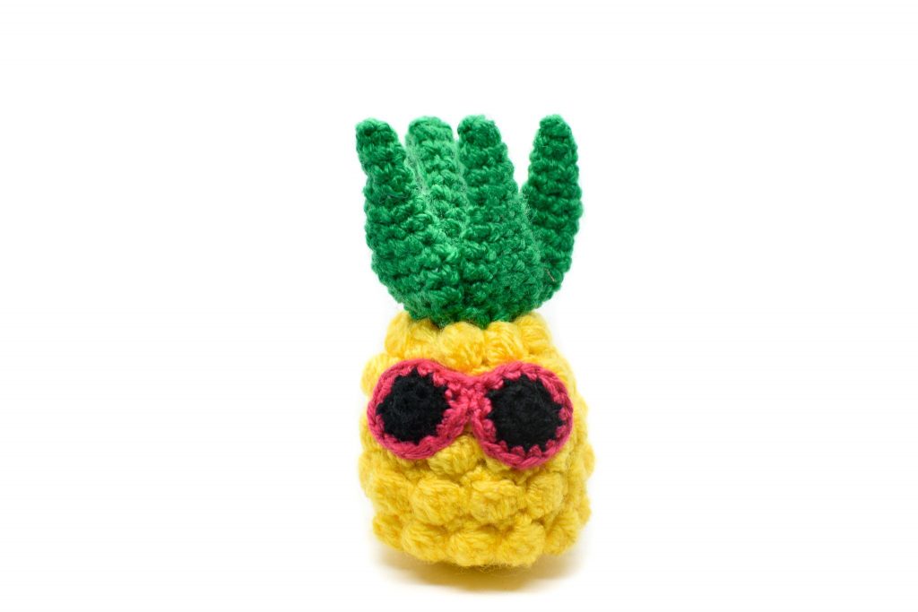 a small crochet pineapple toy with pink sunglasses against a white background