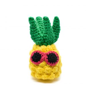 a small crochet pineapple toy with pink sunglasses against a white background