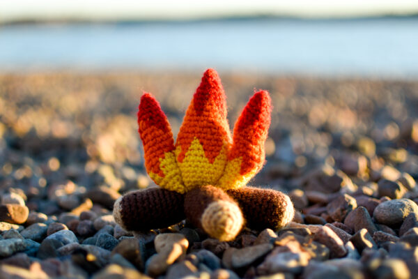a small plush crochet campfire sitting on a rocky beach at sunset