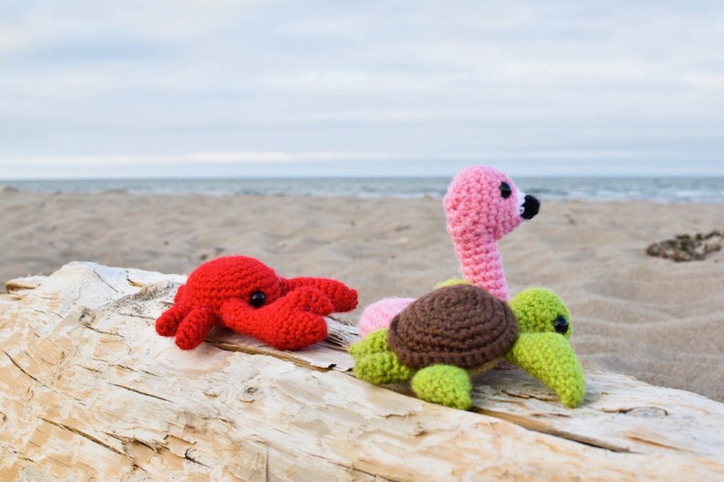 three small crochet toys sitting on a log at the beach - a flamingo floatie, a crab, and a sea turtle