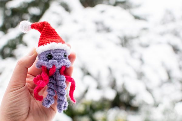 a small purple and pink crochet jellyfish with a Santa Hat, held up against a snowy background