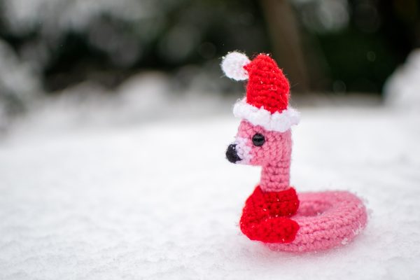a small crochet flamingo floatie with a Santa Hat and red scarf, sitting on snowy ground