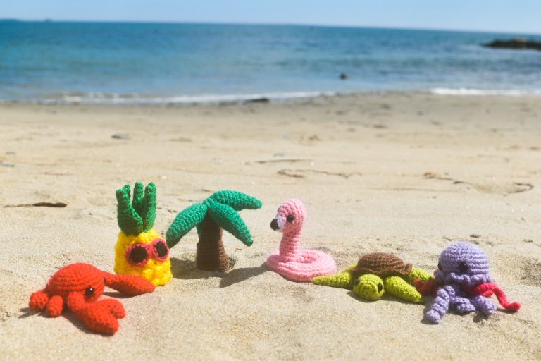 6 small crochet figures sitting on a beach: a crab, a pineapple with pink sunglasses, a palm tree, a flamingo floatie, a sea turtle, and a purple jellyfish