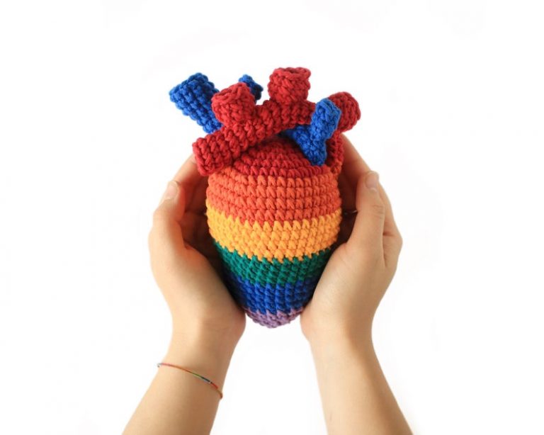 two hands holding a crocheted anatomical heart made in striped rainbow colours