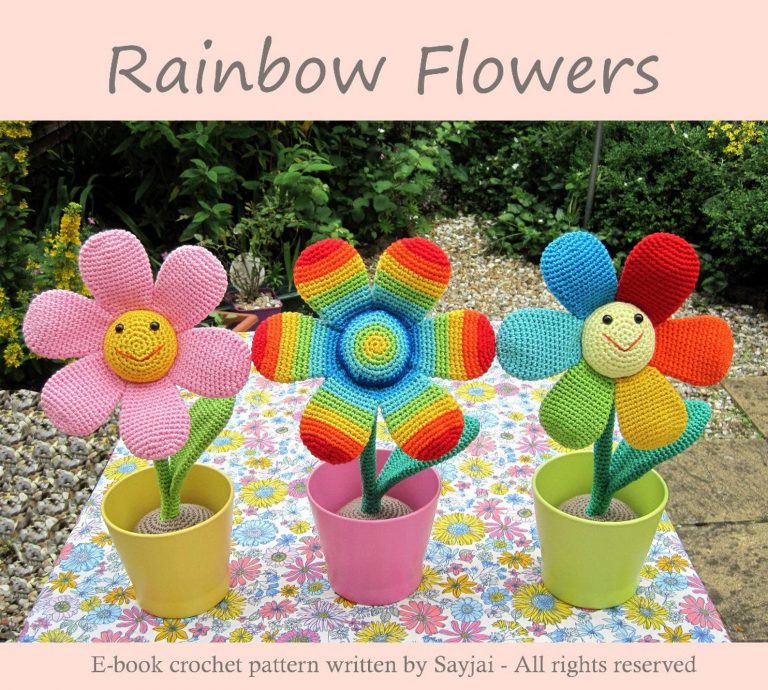three cute crocheted flowers placed into pots. One has a yellow middle with a happy face and pink petals. The second one has rainbow coloured stripes in the middle and petals. The Third one has a yellow middle with a happy face, and solid rainbow coloured petals.
