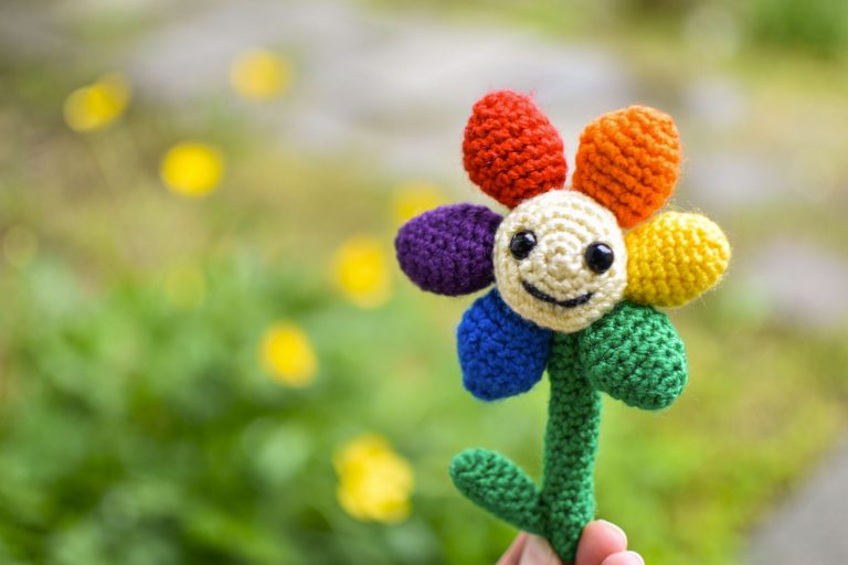 a small crochet flower with a light yellow middle, black plastic eyes and a smiley face. There are 6 solid-coloured petals arranged in rainbow order: red, orange, yellow, green, blue, and purple. The flower is being held up against a blurry background of grass and yellow flowers