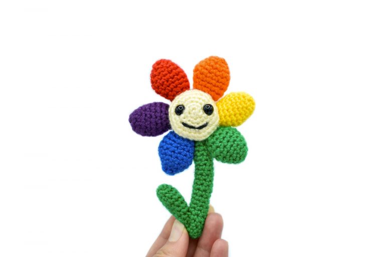 a small crochet flower with a light yellow middle, black plastic eyes and a smiley face. There are 6 solid-coloured petals arranged in rainbow order: red, orange, yellow, green, blue, and purple. The flower is being held up against a white background