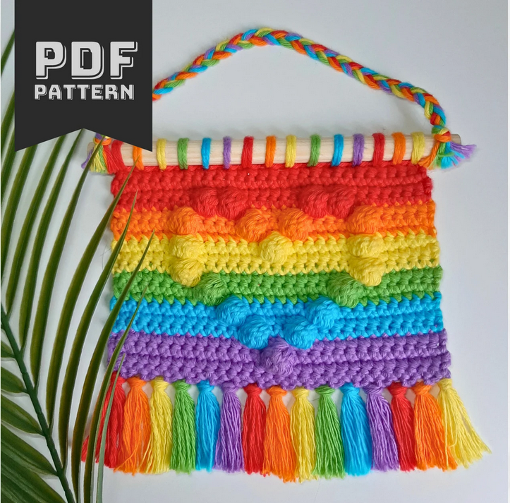 A small crochet wall hanging made in rainbow colours with a heart shape motif formed by bobble stitches. There are rainbow tassels at the bottom.
