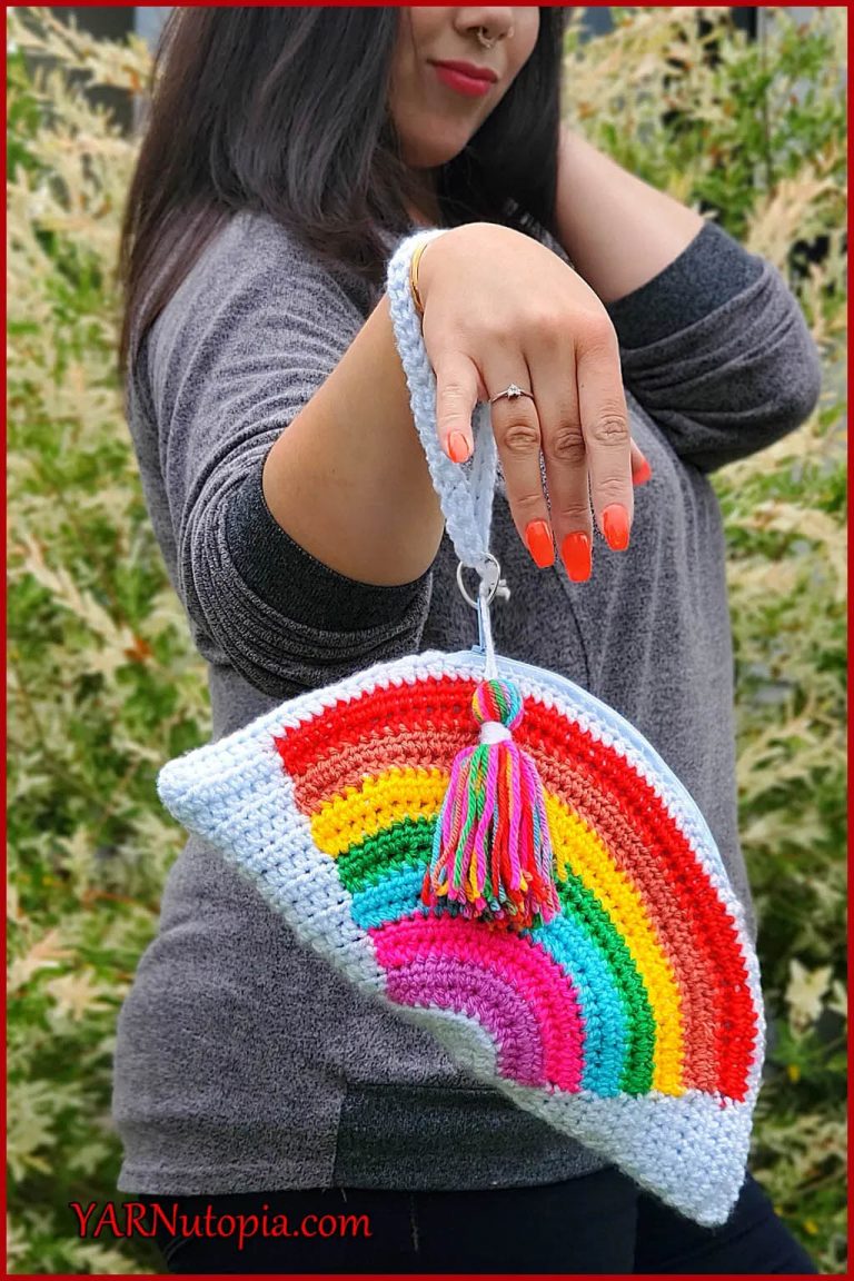 A woman with a crochet clutch bag hanging off her wrist. The clutch bag is white with a rainbow on it, and the zipper has a rainbow coloured tassel. The bag is approx 1 foot long at its widest spot.