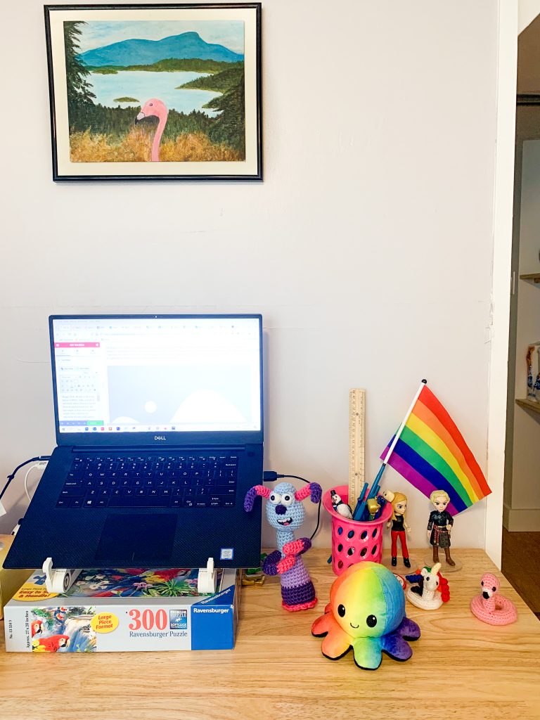 a picture of a home office desk set up. There is an open laptop computer on the desk, and next to it are several items including: a pink pen holder with some pens and a ruler, a small rainbow Pride flag, two toy figurines of Buffy the Vampire Slayer and Brienne from Game of Thrones, a small crochet unicorn floatie and flamingo floatie, a crochet Lu-la from the Shaun the Sheep movie, and a plush rainbow coloured octopus. Above the desk is a painting of an ocean and mountain view with a flamingo in front of it.
