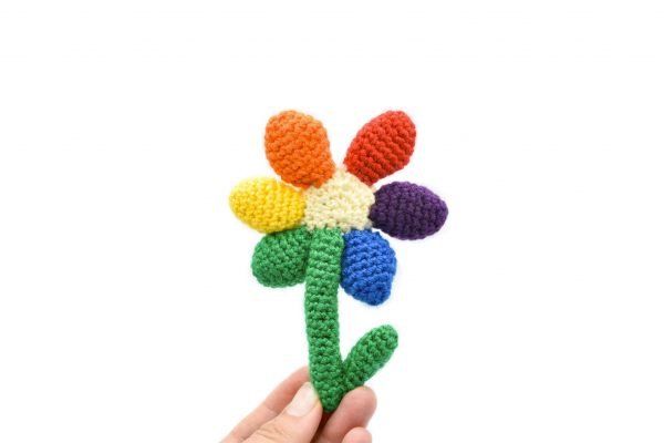 back view of a small crochet flower with a light yellow middle and a green stem with a small leaf. There are 6 solid-coloured petals arranged in rainbow order: red, orange, yellow, green, blue, and purple. The flower is being held up against a white background.