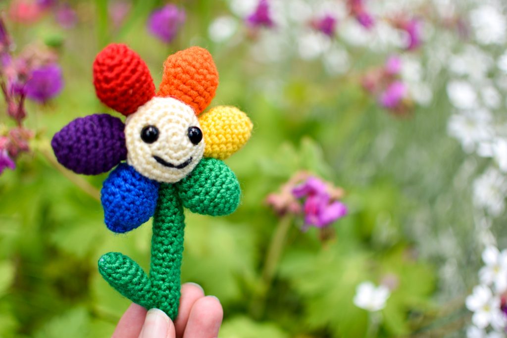 a small crochet flower with a light yellow middle, black plastic eyes and a smiley face. There are 6 solid-coloured petals arranged in rainbow order: red, orange, yellow, green, blue, and purple. The flower is being held up against a background of greenery and pink & white flowers