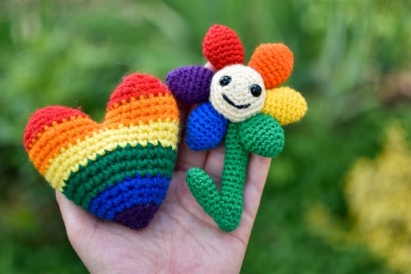 a hand holding a rainbow-coloured crochet heart and a small crochet flower with a light yellow middle, black plastic eyes and a smiley face. There are 6 solid-coloured petals arranged in rainbow order: red, orange, yellow, green, blue, and purple. The flower and heart are being held up against a background of greenery