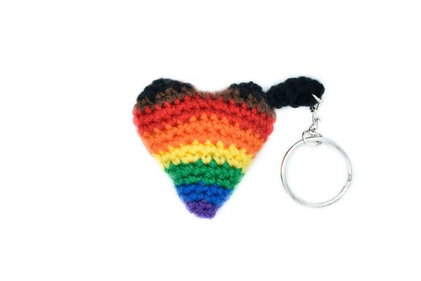 a small crochet heart keychain in the inclusive rainbow pride flag colours against a white background. the inclusive rainbow flag is the regular rainbow + brown and black.