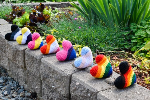 Eight stuffed crochet hearts lined up on a stone wall in a garden. Each heart represents the colours of a different Pride flag.