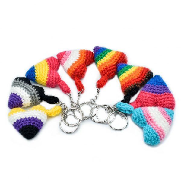 8 small crochet heart keychains with pride colours, arranged in a semi-circle. The pride flags are: Rainbow, Inclusive Rainbow, Trans, Bisexual, Lesbian, Asexual, Pansexual, and Non-Binary flag colours.