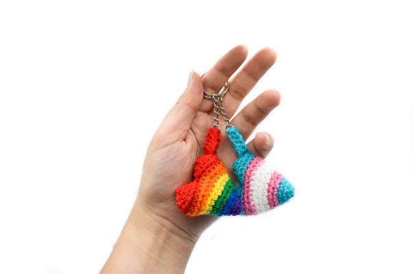 a hand holding two small crochet heart keychains. The keychain rings are over one finger, and both hearts are dangling down into the palm of the hand. One heart is rainbow pride colours, and the other heart is the trans pride flag colours (light blue, light pink, and white)
