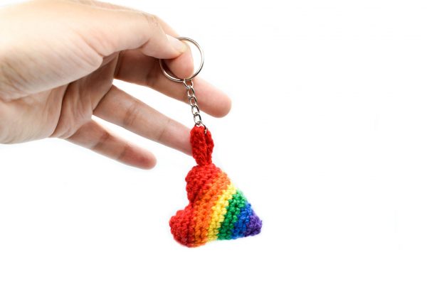 a hand holding a small crochet heart keychain in the rainbow pride flag colours, against a white background. The hand is holding the key ring and dangling the heart down.