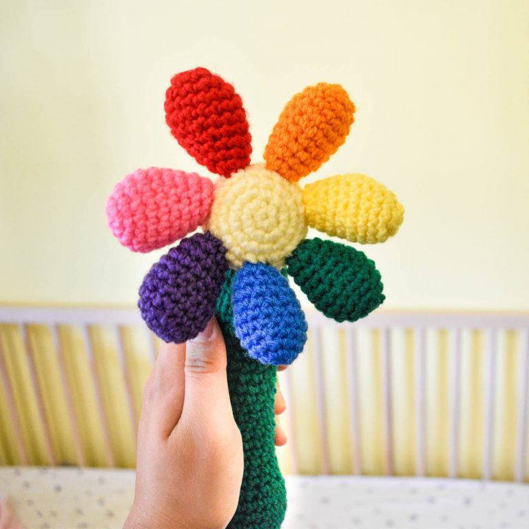 a crochet baby rattle in the shape of a flower. The handle is green, the centre of the flower is light yellow, and there are 7 round petals in red, orange, yellow, green, blue, purple, and pink. The rattle is bring held up in front of a baby crib