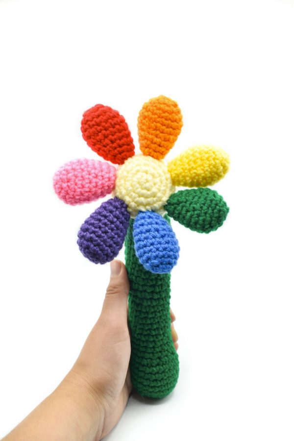 a crochet baby rattle against a white background. The rattle is in the shape of a flower. The handle is green, the centre of the flower is light yellow, and there are 7 round petals in red, orange, yellow, green, blue, purple, and pink.