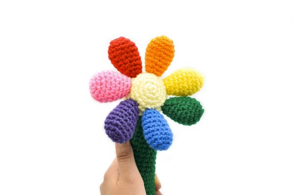 a crochet baby rattle against a white background. The rattle is in the shape of a flower. The handle is green, the centre of the flower is light yellow, and there are 7 round petals in red, orange, yellow, green, blue, purple, and pink.