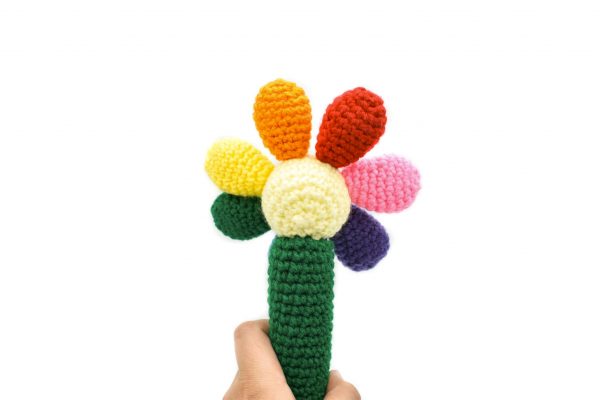 back view of a crochet baby rattle against a white background. The rattle is in the shape of a flower. The handle is green, the centre of the flower is light yellow, and there are 7 round petals in red, orange, yellow, green, blue, purple, and pink.