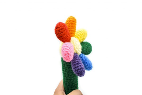side view of a crochet baby rattle against a white background. The rattle is in the shape of a flower. The handle is green, the centre of the flower is light yellow, and there are 7 round petals in red, orange, yellow, green, blue, purple, and pink.