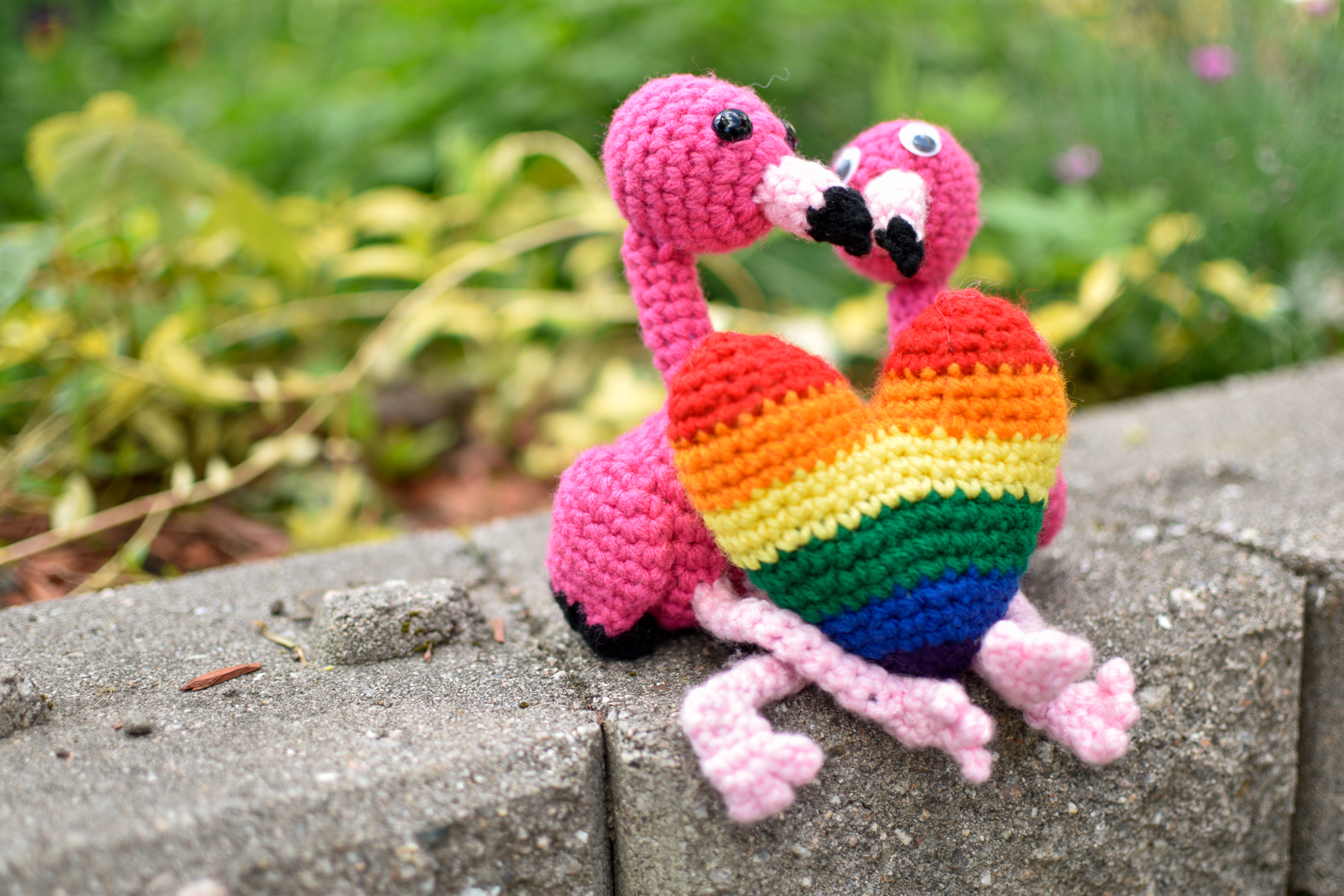 Two stuffed crochet flamingos sitting next to each other on a stone wall with a crochet rainbow heart between then.