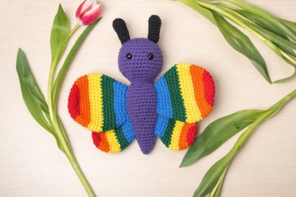 a stuffed crochet butterfly toy with a purple body, black antennae, and rainbow coloured wings. The butterfly is lying on a beige background surrounded by pink tulips