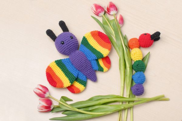 a stuffed crochet butterfly toy lying on a beige background next to a stuffed crochet caterpillar toy. They are surrounded by pink tulips. The butterfly has a purple body, black antennae, and rainbow coloured wings. The caterpillar is also made in rainbow colours, with the head and each body segment a different colour of the rainbow