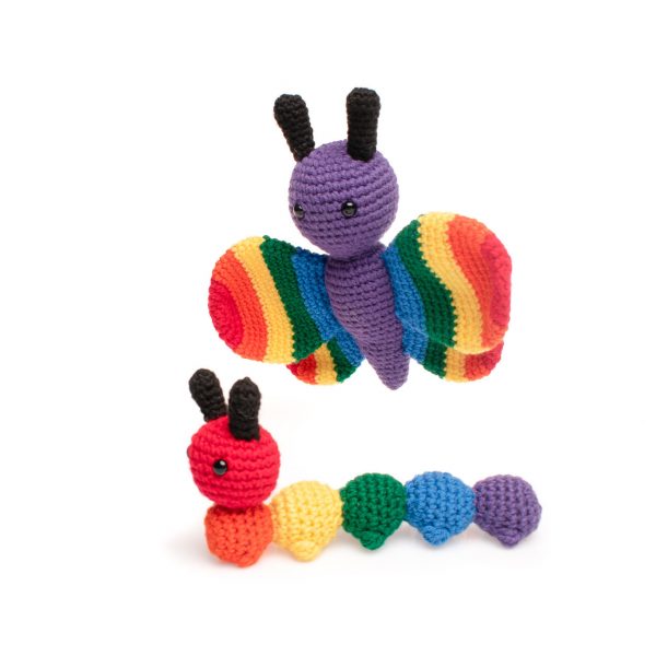 a stuffed crochet butterfly toy hovering over a stuffed crochet caterpillar toy. The butterfly has a purple body, black antennae, and rainbow coloured wings. The caterpillar is also made in rainbow colours, with the head and each body segment a different colour of the rainbow