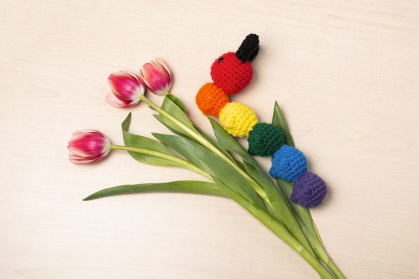 a crochet stuffed caterpillar made in rainbow colours, with the head and each body segment a different colour of the rainbow. The caterpillar is lying on a beige background and is climbing a set of pink tulips