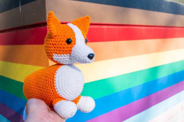 a crochet stuffed corgi dog being held up in front of a wall painted in Pride flag colours