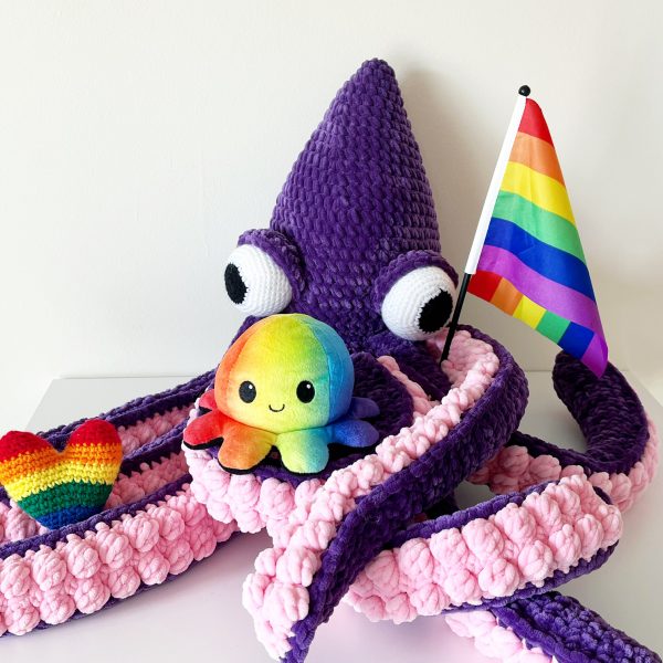 a large crochet kraken made out of chunky yarn. The body is purple and the underside of the tentacles are light pink. The kraken is holding a stuffed rainbow heart, rainbow octopus, and a Pride flag in its tentacles