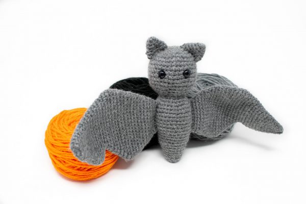 a stuffed crochet grey bat posed in front of balls of orange and black yarn