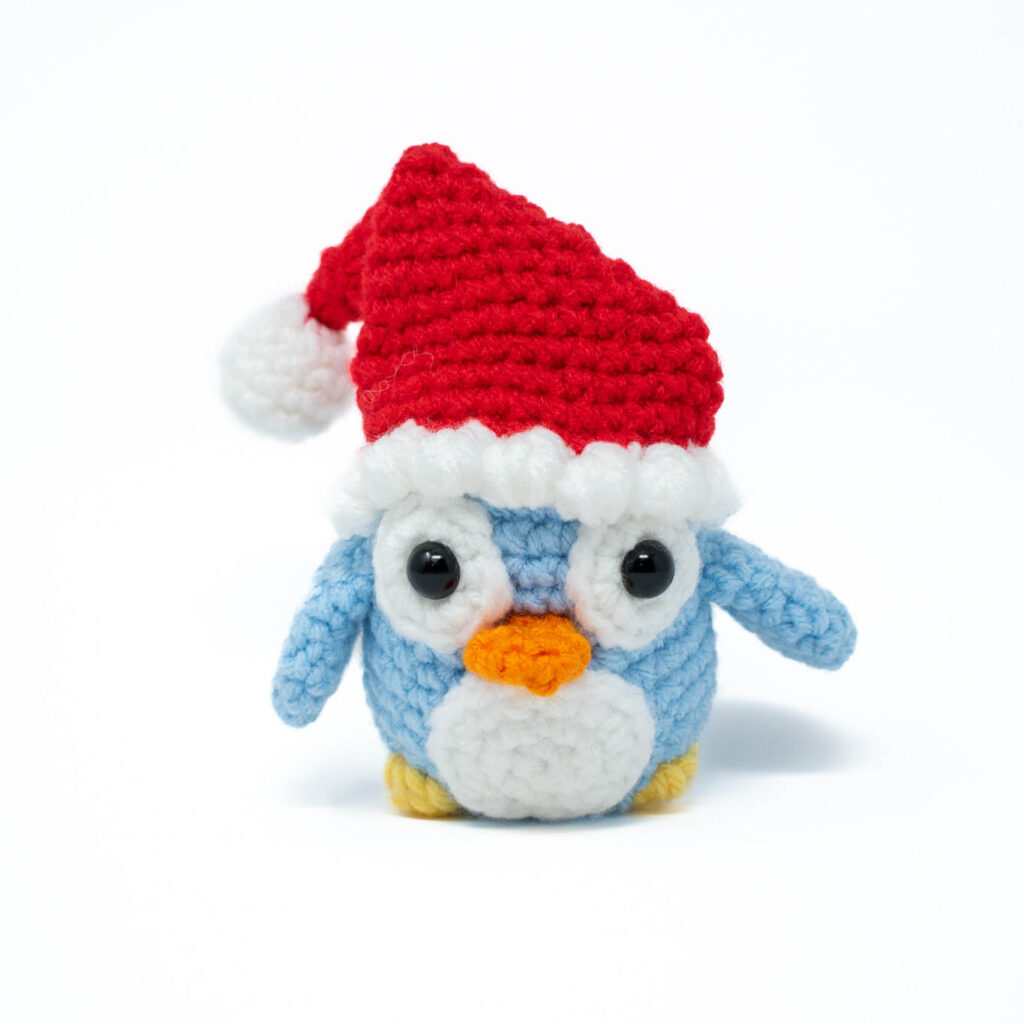 a light blue crochet stuffed penguin with a Santa hat