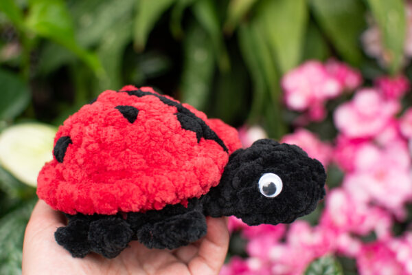 a crochet ladybug toy made from soft chunky chenille yarn, being held up in front of some pink flowers