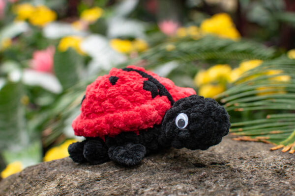 a crochet ladybug toy made from soft chunky chenille yarn, sitting on a rock in front of some yellow and pink flowers