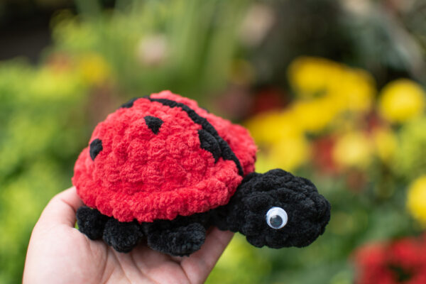 a crochet ladybug toy made from soft chunky chenille yarn, being held up in front of some yellow flowers