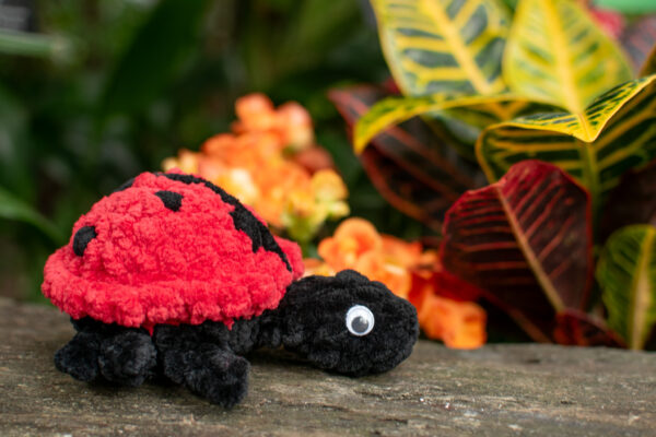 a crochet ladybug toy made from soft chunky chenille yarn, sitting on a log in front of some plants