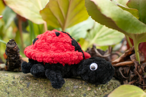 a crochet ladybug toy made from soft chunky chenille yarn, sitting on a rock under some leaves